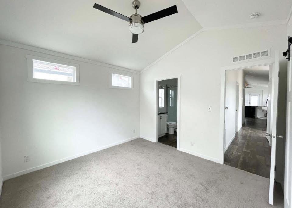 Bright, empty bedroom with white walls and carpet. Features a ceiling fan, two high windows, adjoining bathroom, and hallway leading to a kitchen.