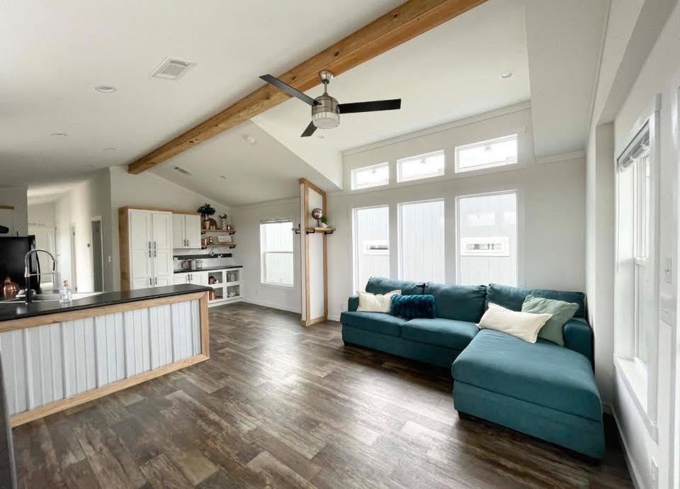 Spacious living room with high ceilings, wooden beams, and large windows. Features a teal sectional sofa, ceiling fan, and rustic hardwood floors.