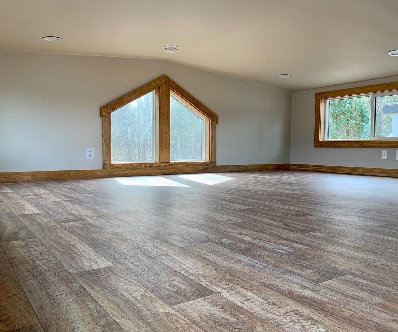 Cozy room with light wood flooring, vaulted ceiling, and two large triangular windows. Sunlight streams in, creating a warm and inviting atmosphere.