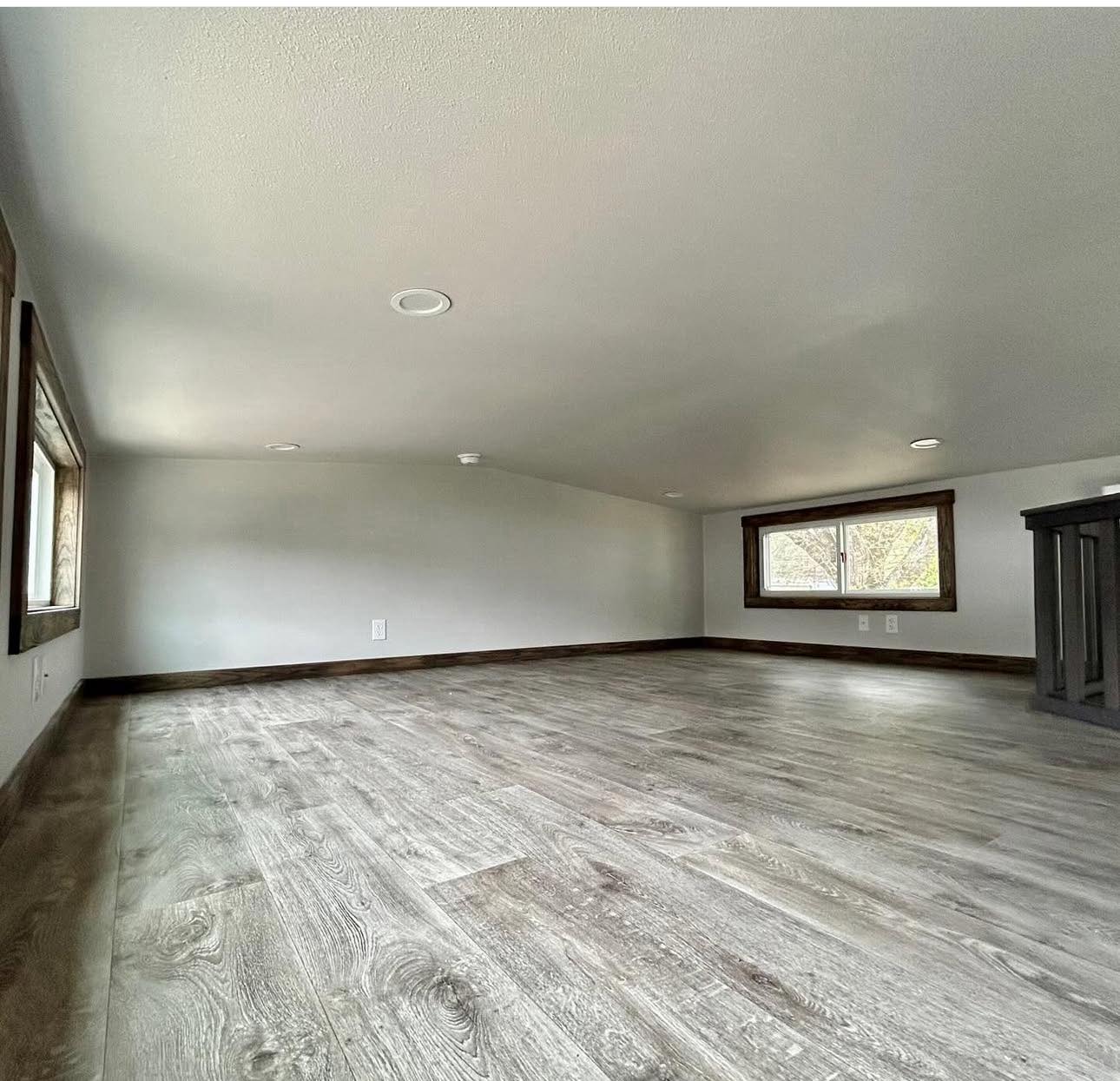 A minimalist attic room with light gray walls and wood-look laminate flooring. Two small windows with wooden frames provide natural light, creating a serene ambiance.