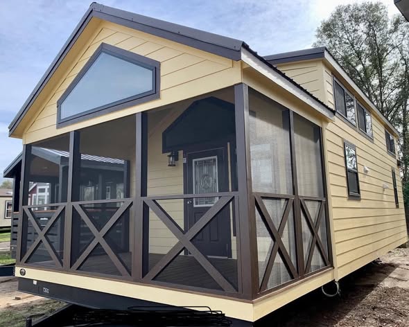 Tiny house with yellow siding and a brown screened porch. A welcoming doorway is visible. The design is cozy and modern, set against a backdrop of trees.