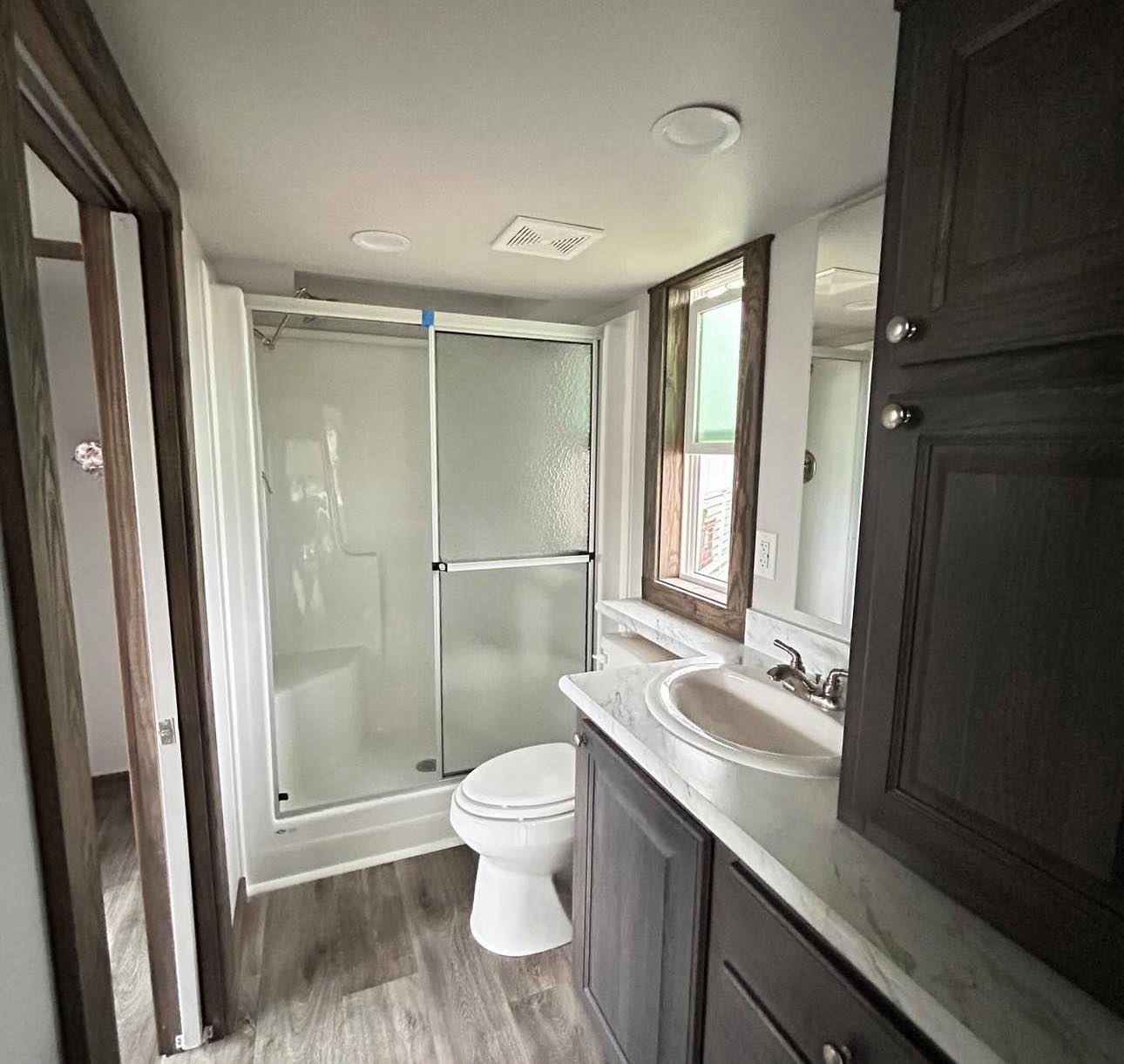 Compact bathroom with wood flooring, featuring a shower with sliding glass doors, a white toilet, marble countertop, and dark wood cabinets. Bright and clean.