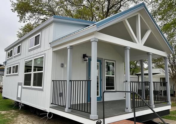 A modern tiny house with a white exterior, blue trim, and a spacious porch. Large windows provide light, creating an inviting and cozy atmosphere.