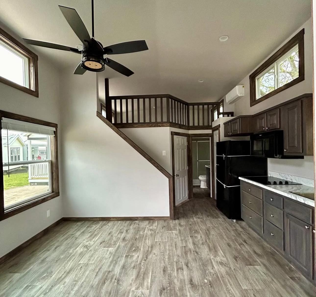 Modern tiny house interior with wood floors, a kitchen on the right with dark cabinets, loft with railing above, large windows, and a ceiling fan. Cozy and bright ambiance.
