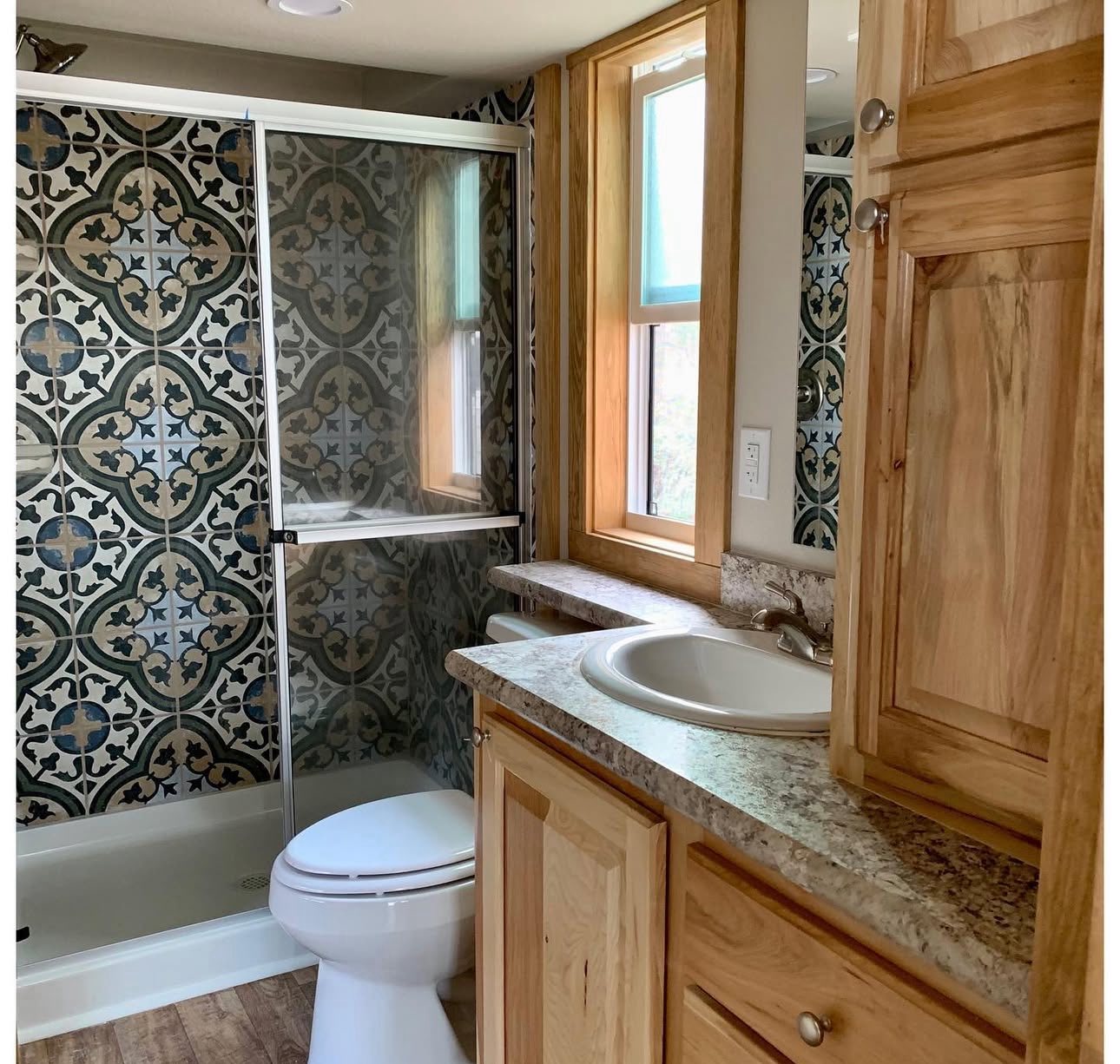 Bathroom with patterned tile shower, wooden cabinetry, granite countertop, sink, and toilet. Bright window adds natural light. Cozy and modern.