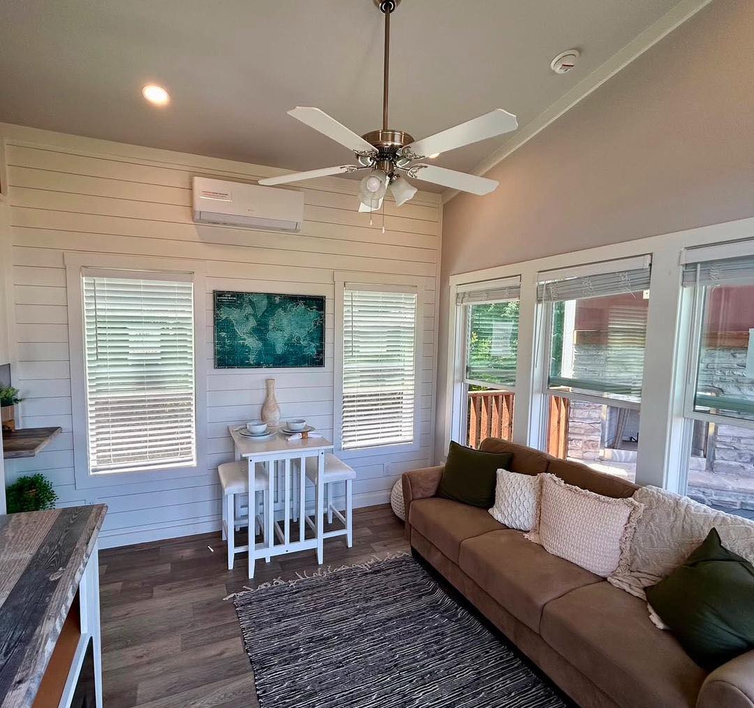 Cozy living room with a beige sofa adorned with green and white pillows, a small dining table with vases, wooden floor, window blinds, and a ceiling fan.
