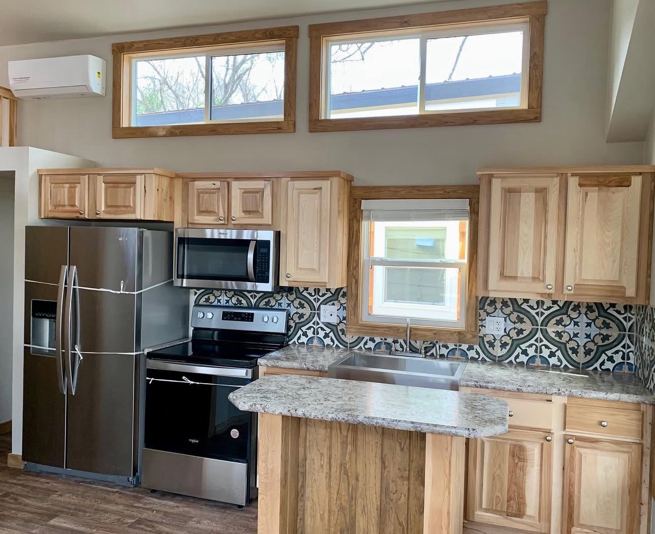 Bright kitchen with wooden cabinets, stainless steel appliances, stone countertops, and patterned tile backsplash. Large windows add natural light. Cozy, modern feel.
