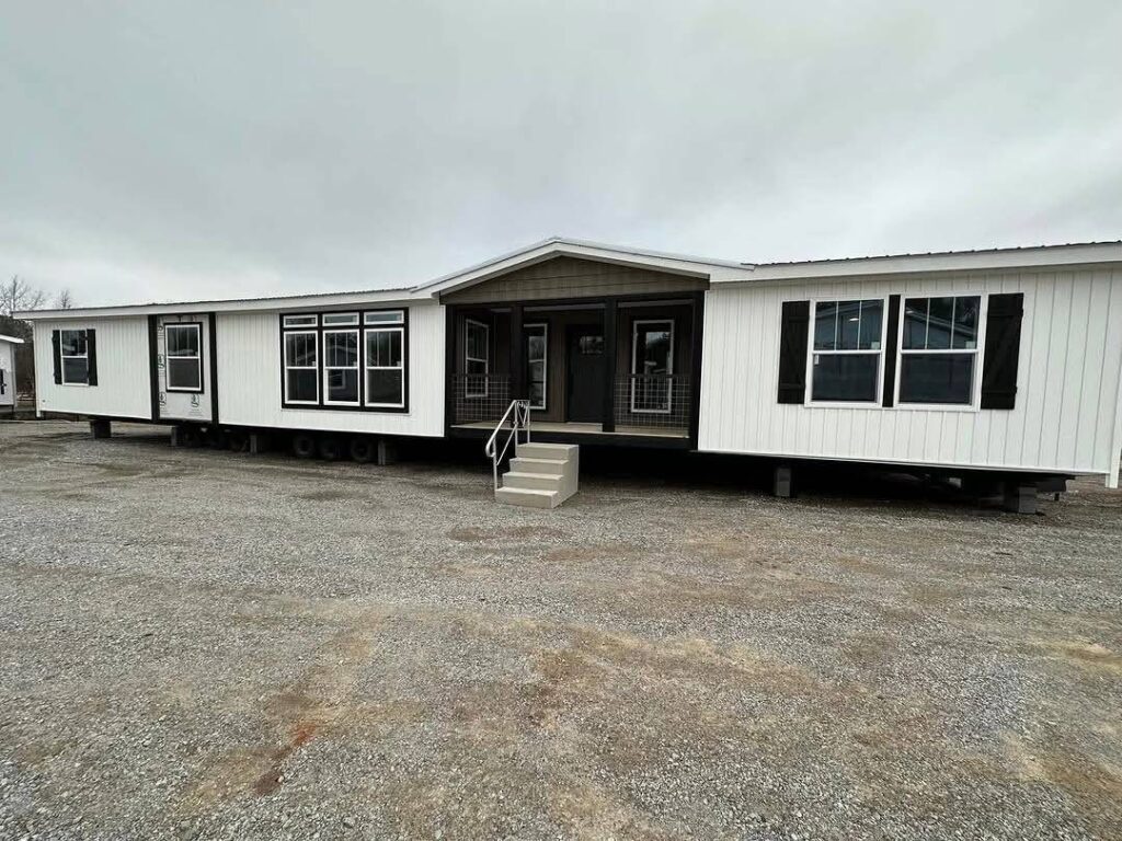 A spacious, double-wide mobile home with white siding and black trim is set on a gravel lot. It features multiple windows and a small stoop with steps leading to a black front door, under a cloudy sky.