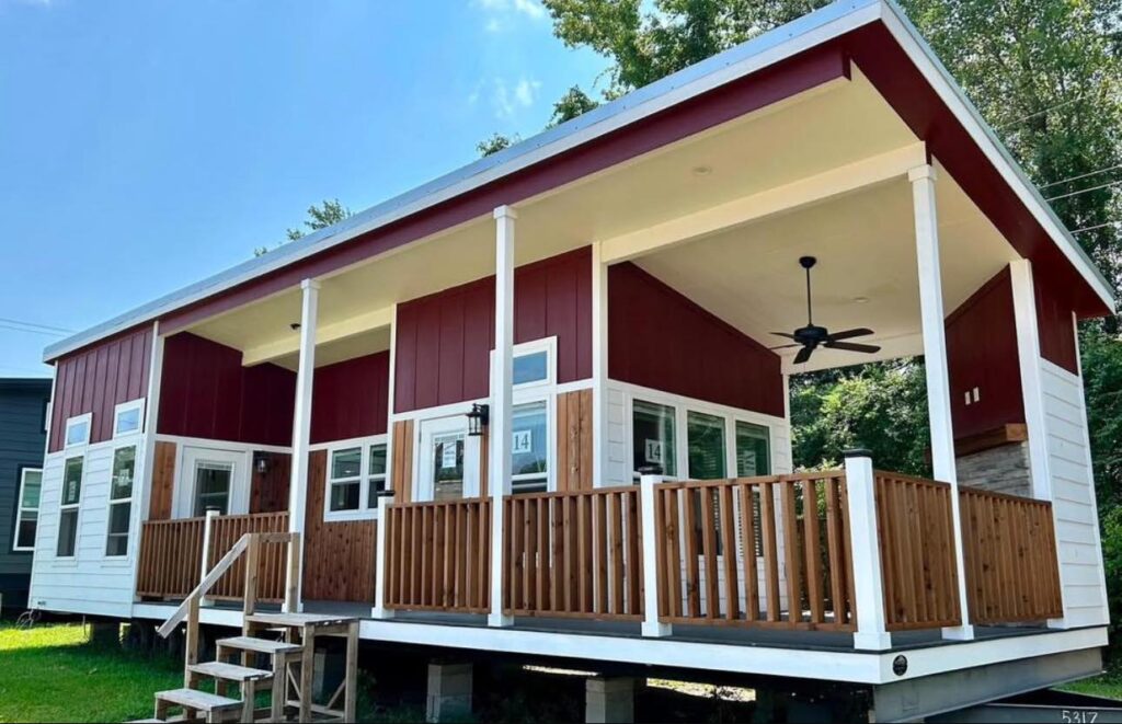 Modern tiny house with a red and white exterior, large windows, and a wooden porch. Elevated on stilts, it features a ceiling fan and staircase, set against a blue sky.