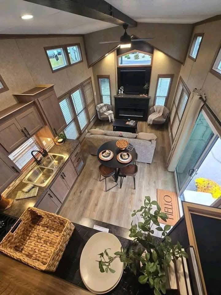 A cozy living room with high ceilings, viewed from above. Features include a grey sofa, armchairs, a round dining table set for two, and a TV above a fireplace. Sunlight filters through large windows, illuminating plants and a welcoming atmosphere.