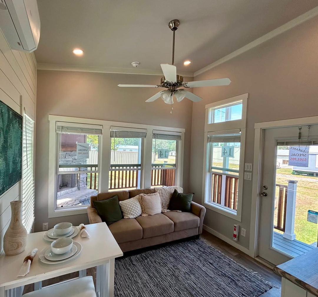 A cozy living room with neutral tones features a beige sofa adorned with cushions. Large windows brighten the space, which includes a small dining table set for two. A ceiling fan adds comfort, while natural light creates an inviting atmosphere.
