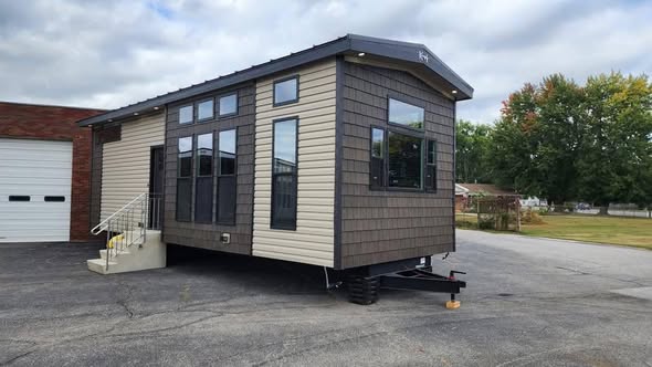 A modern tiny house with dark siding and large windows sits on a paved lot. It has a small porch with steps, a cloudy sky, and trees in the background.