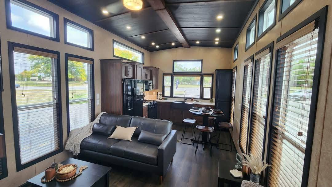 Modern tiny home interior featuring a sleek black sofa, wood finishes, large windows, and a compact kitchen. Bright, cozy, and inviting space.