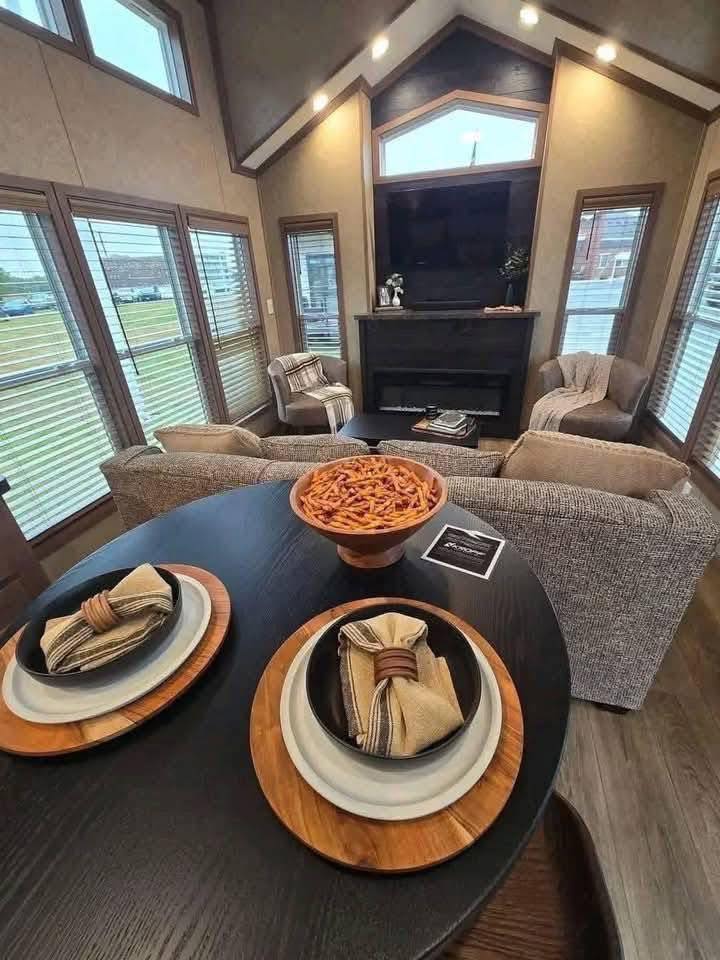 Cozy living room with modern decor. A round table holds two place settings with beige napkins and a bowl of snacks. Soft lighting and large windows create a warm, inviting atmosphere.