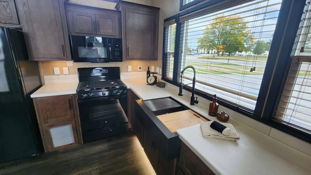 Modern kitchen with dark wood cabinets, black appliances, and large windows. Sunlight illuminates a sleek sink area, evoking warmth and elegance.