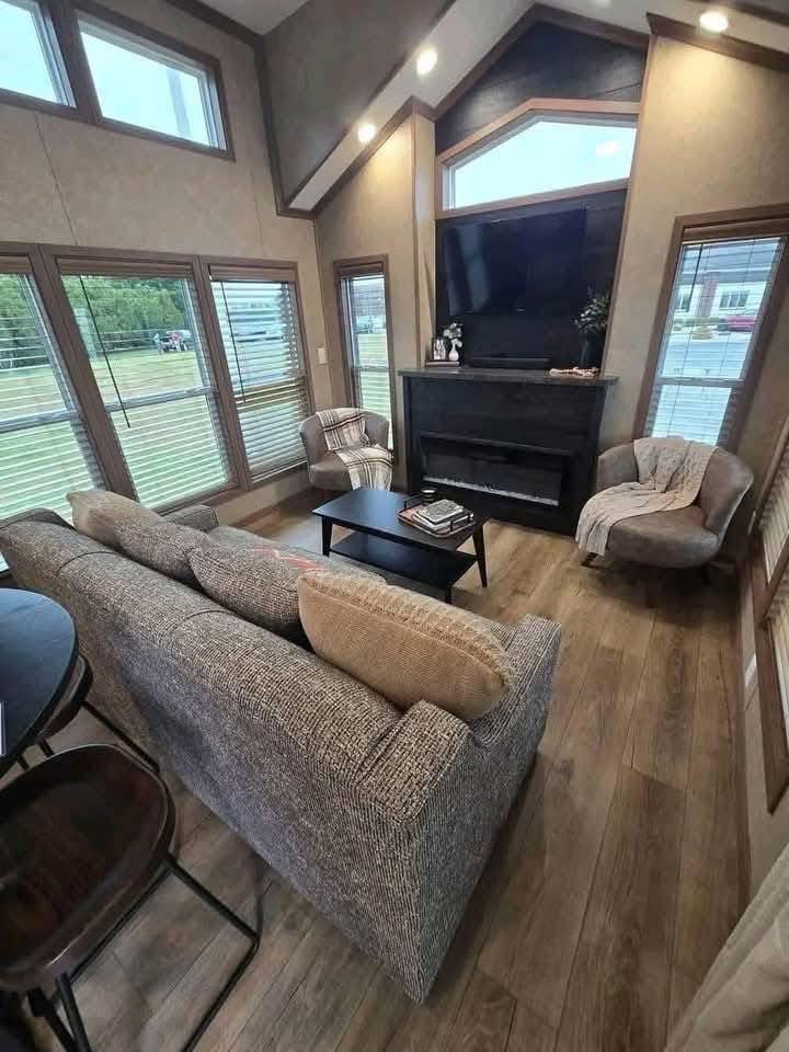 Cozy living room with high ceilings and large windows, featuring a gray couch, two armchairs, a black coffee table, and a mounted TV above a fireplace. Warm tones create a welcoming atmosphere.