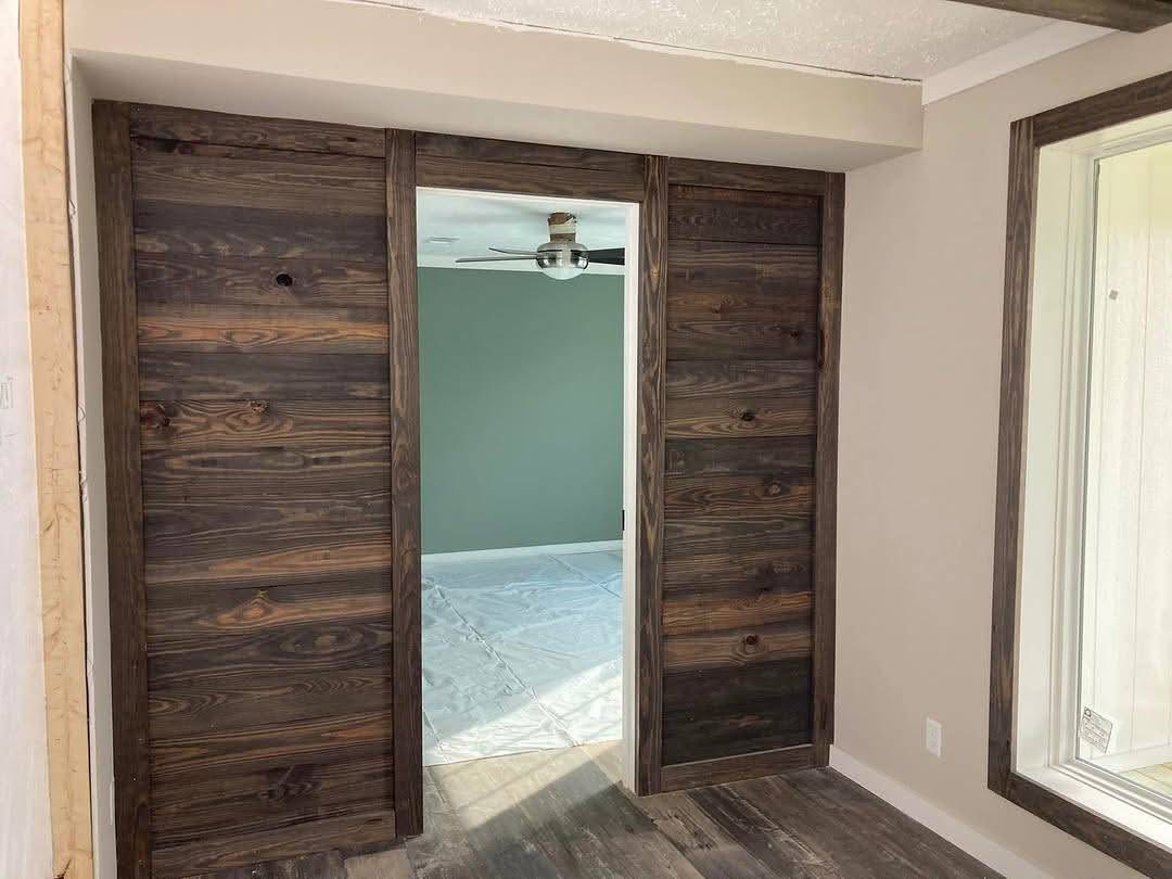 Rustic sliding barn doors with dark wood grain frame an opening to a room with green walls and a ceiling fan. The room has plastic-covered floors.