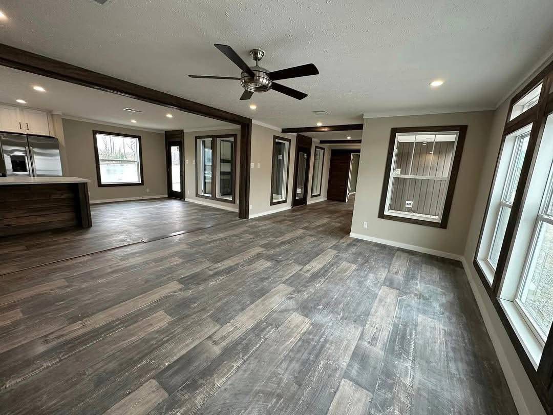 Spacious modern living area with rustic wood flooring, large windows, and ceiling fan; open layout features kitchen with stainless steel appliances.