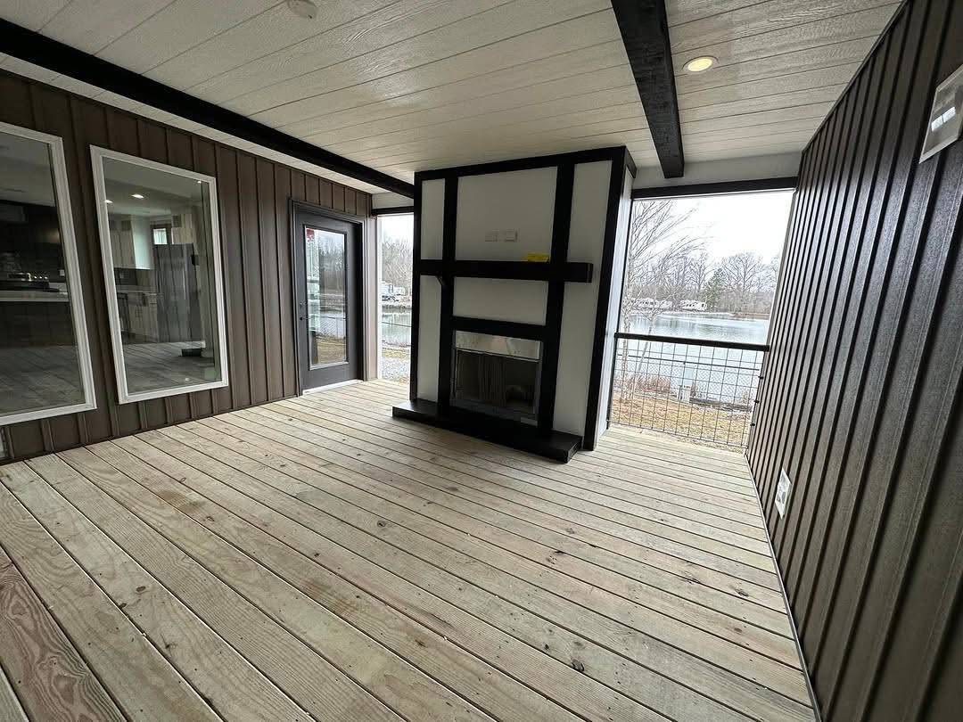 Covered patio with light wood deck and dark paneling, featuring a modern black and white fireplace. Large windows show a lakeside view.