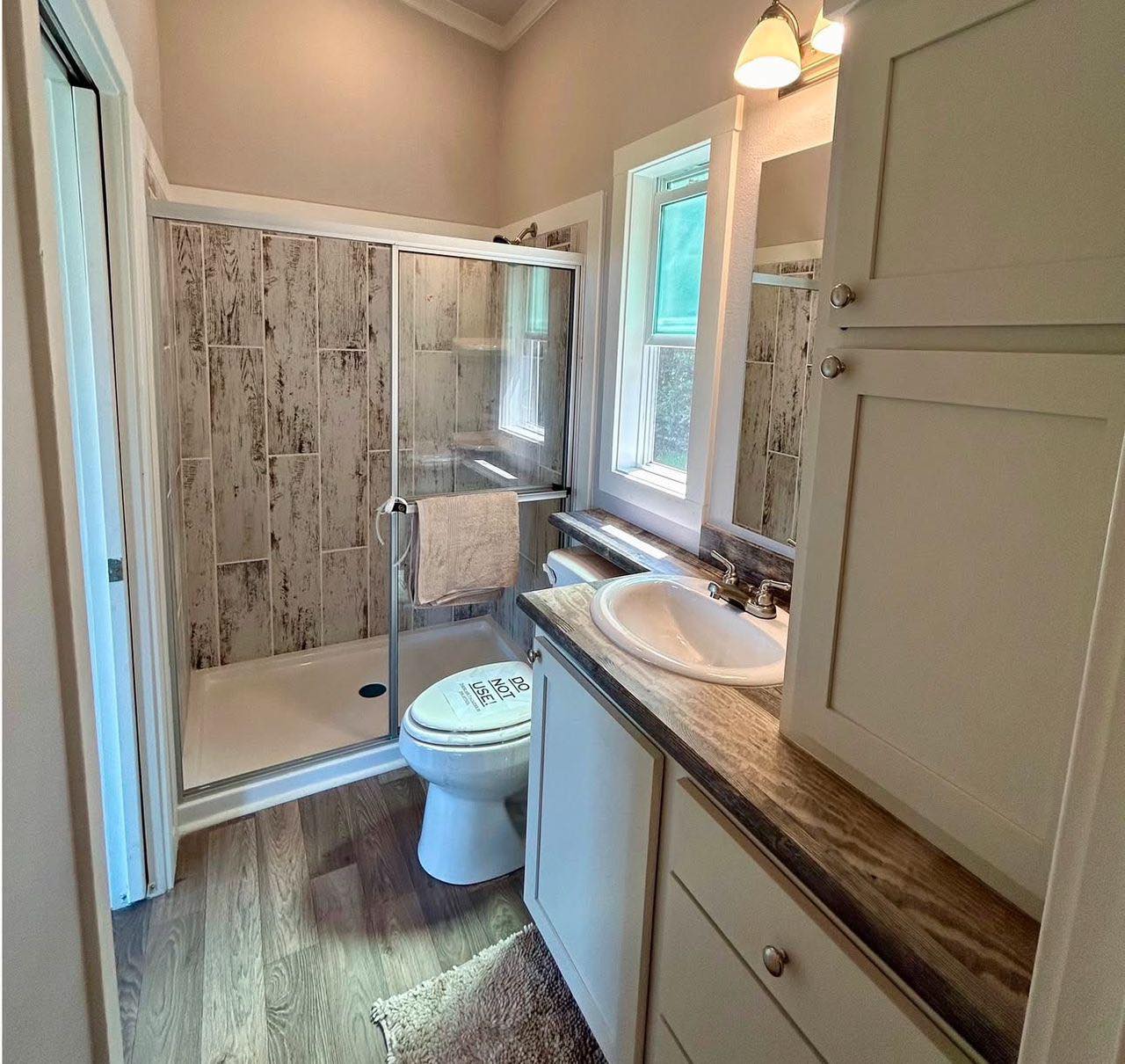 A compact bathroom with a rustic wood-tiled shower, glass doors, and natural light. White vanity with a brown countertop and modern fixtures.