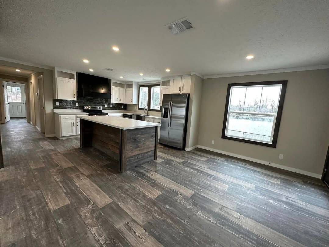 Spacious modern kitchen with a central island, light wood floors, and stainless steel appliances. Large windows offer a view of trees and water.