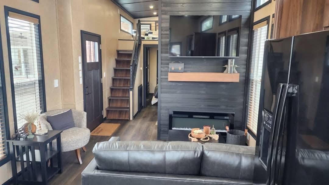 A modern living room with a black leather sofa facing a large wall-mounted TV above a fireplace. Wooden stairs lead to a loft. The room features large windows, light gray walls, and dark floors, creating a cozy and contemporary feel.