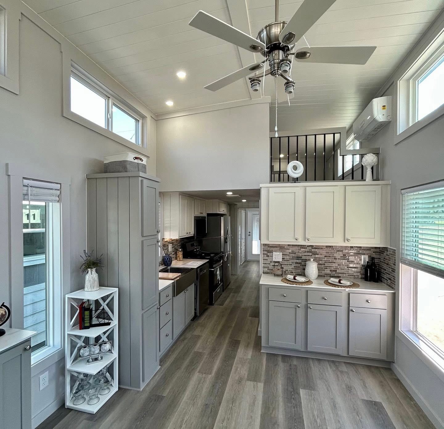Modern tiny house kitchen with light wood floors, gray cabinets, and stainless steel appliances. Loft area above. Bright, airy, and minimalist design.
