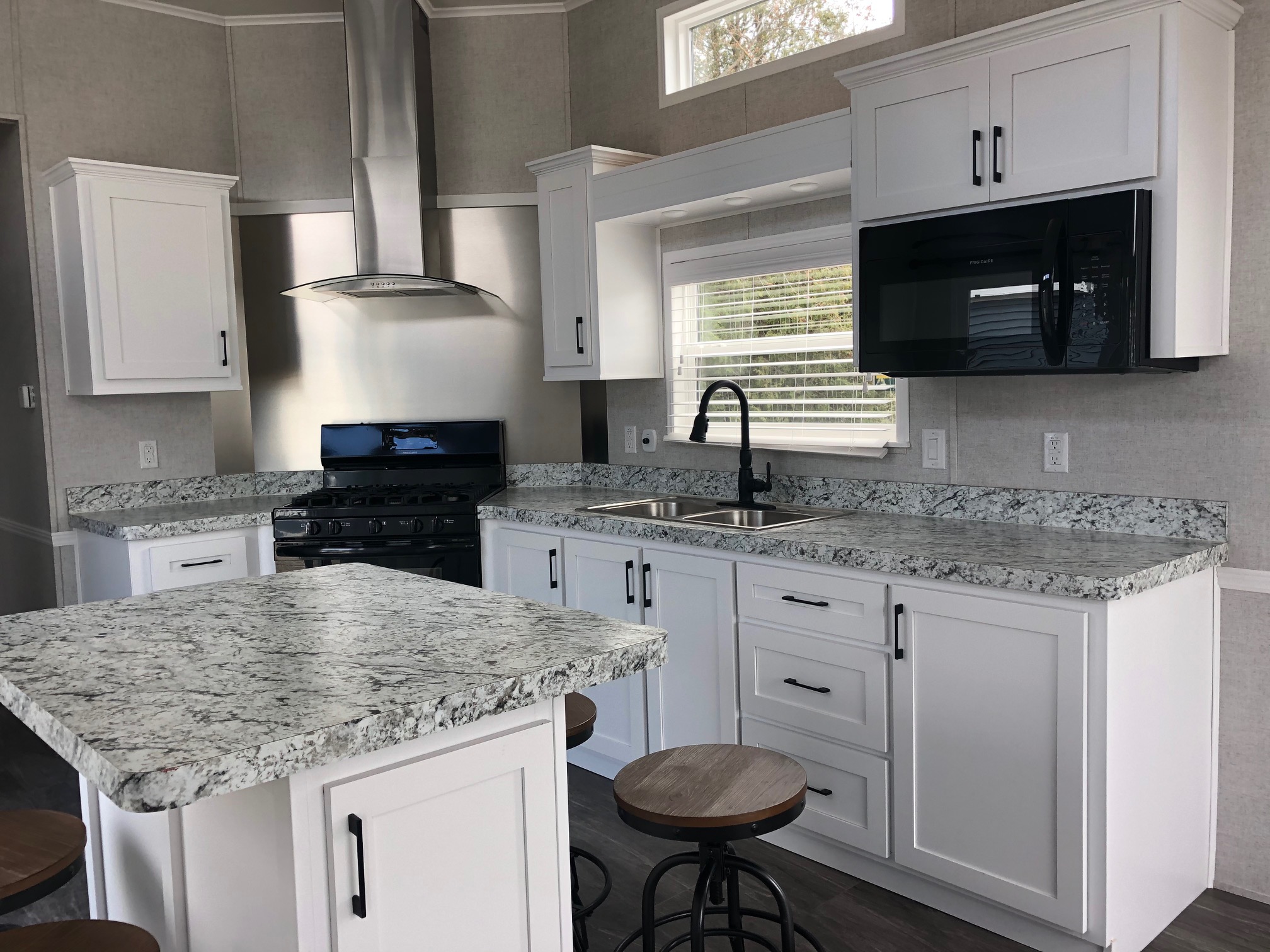Modern kitchen with white cabinets, granite countertops, and stainless steel appliances. Features island with bar stools and a large window. Bright and airy.