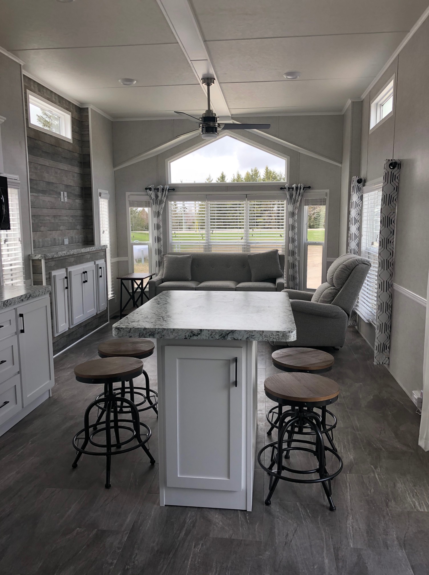 Spacious, modern kitchen with white cabinets, gray countertops, and bar stools. Cozy living area with a large window, couch, and armchair. Bright and inviting.