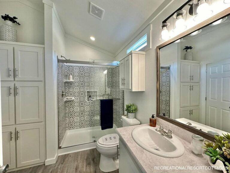 Compact modern farmhouse bathroom in recreational resort cottage featuring bright white shaker cabinets with brushed nickel hardware, large framed vanity mirror with integrated LED lighting, spacious quartz countertop in soft pink-beige tones, oval undermount sink with chrome faucet, patterned gray geometric subway tile shower surround with glass enclosure, walk-in tiled shower with rainfall head and niche shelving, white toilet, luxury vinyl plank flooring in warm gray, vaulted ceiling with recessed lights, high clerestory window for natural light, potted greenery, navy blue towel, decorative vases, and serene spa-like ambiance perfect for cozy cottage retreats or tiny home living.