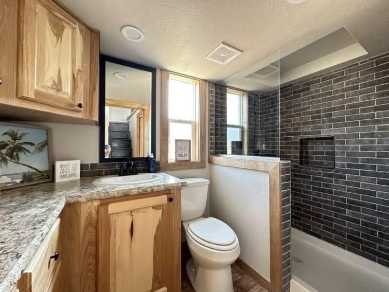 Compact bathroom with light wood cabinets and granite countertop. Features a white toilet, dark brick tile shower, two windows, and a tropical art piece.
