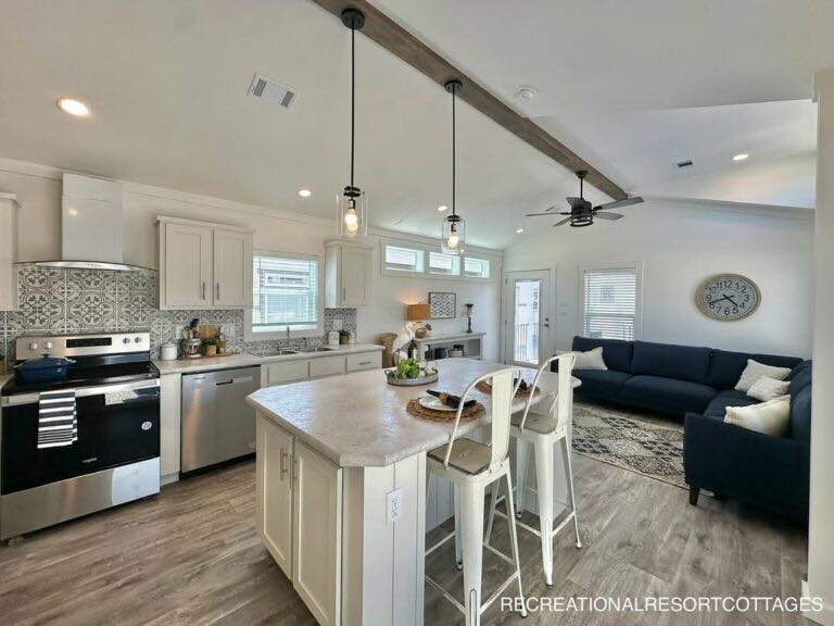 Spacious open-concept modern farmhouse kitchen and living area in a recreational resort cottage featuring vaulted ceiling with exposed rustic wooden beams, black pendant lights over large quartz-top island with white bar stools, stainless steel appliances including gas range and dishwasher, patterned gray subway tile backsplash, shaker white cabinets, cozy navy blue sectional sofa with neutral throw pillows, ceiling fan, luxury vinyl plank flooring, high clerestory windows and large sliding doors for abundant natural light, fresh fruit bowl, potted plants, wall clock, and inviting family-friendly layout perfect for vacation retreats, tiny home living, or cozy cottage getaways.