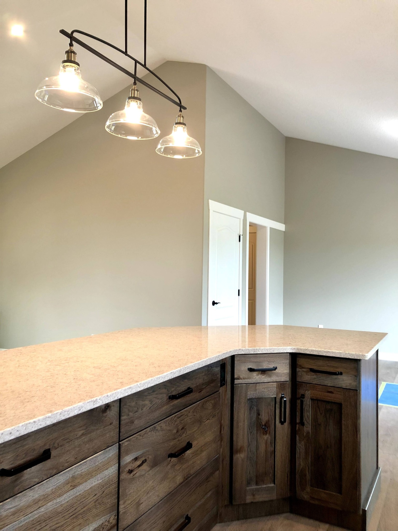 Kitchen with wooden cabinetry, light beige countertop, and three pendant lights. Walls are light gray with high ceilings, creating a spacious feel.