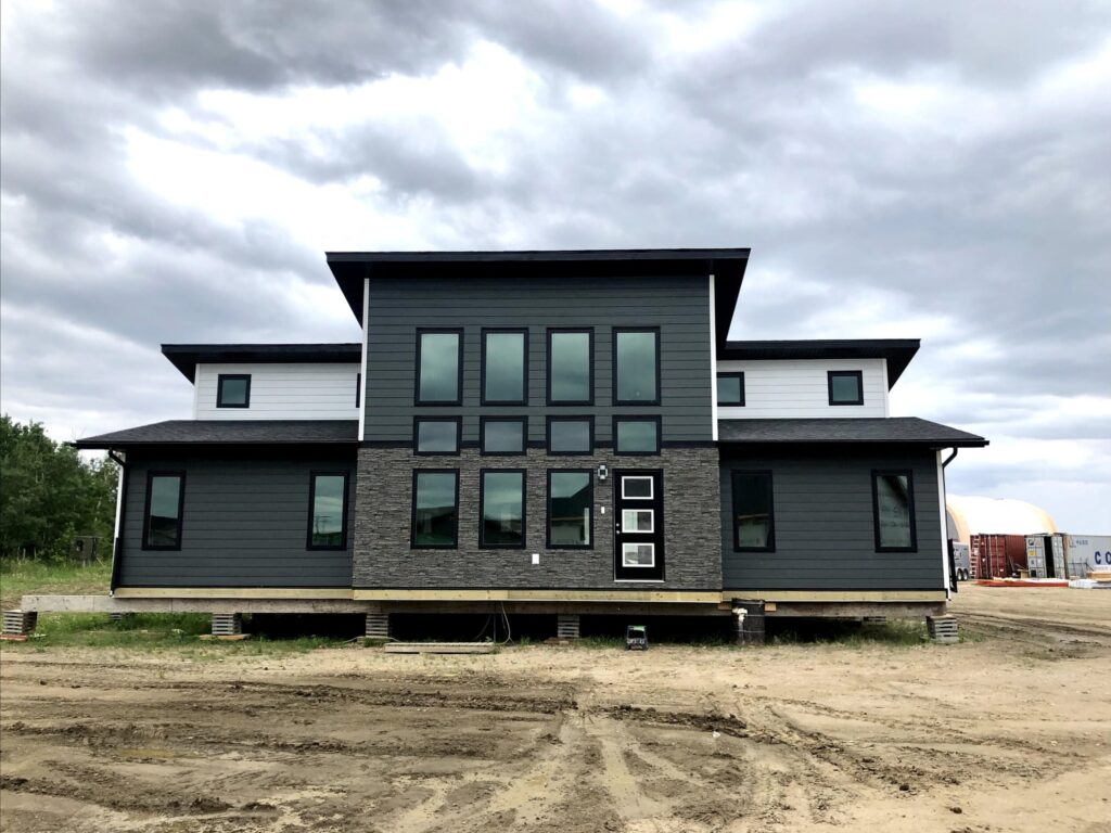Modern gray house with large windows and horizontal siding set against a cloudy sky. The structure is elevated on stilts above bare, dirt ground.