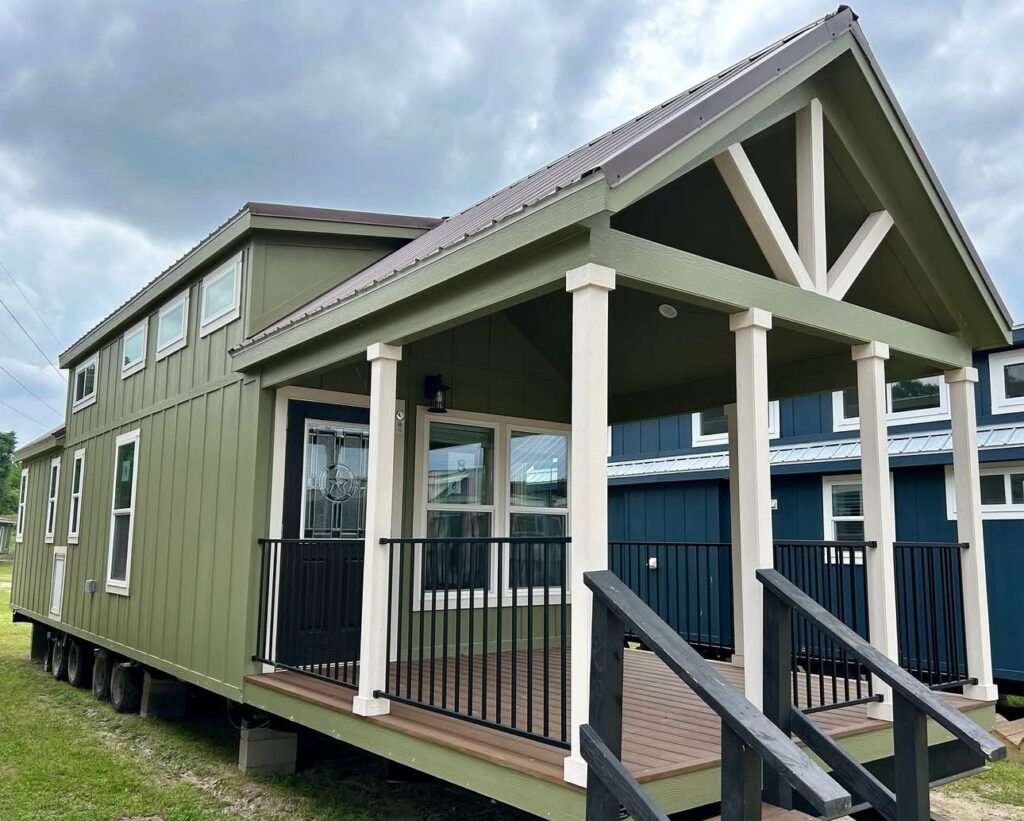 A small, olive-green tiny house with a covered porch and black railings sits against a cloudy sky. The design features large windows and a pitched roof.