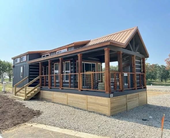 Modern tiny house with a wooden porch, large windows, and a gable roof. It sits on gravel, surrounded by trees under a clear blue sky. Cozy and inviting.