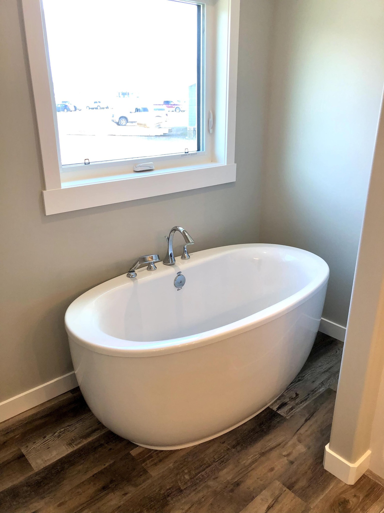 Freestanding white bathtub in a corner near a window, with modern fixtures. Warm wooden floor and neutral walls create a calm, inviting atmosphere.