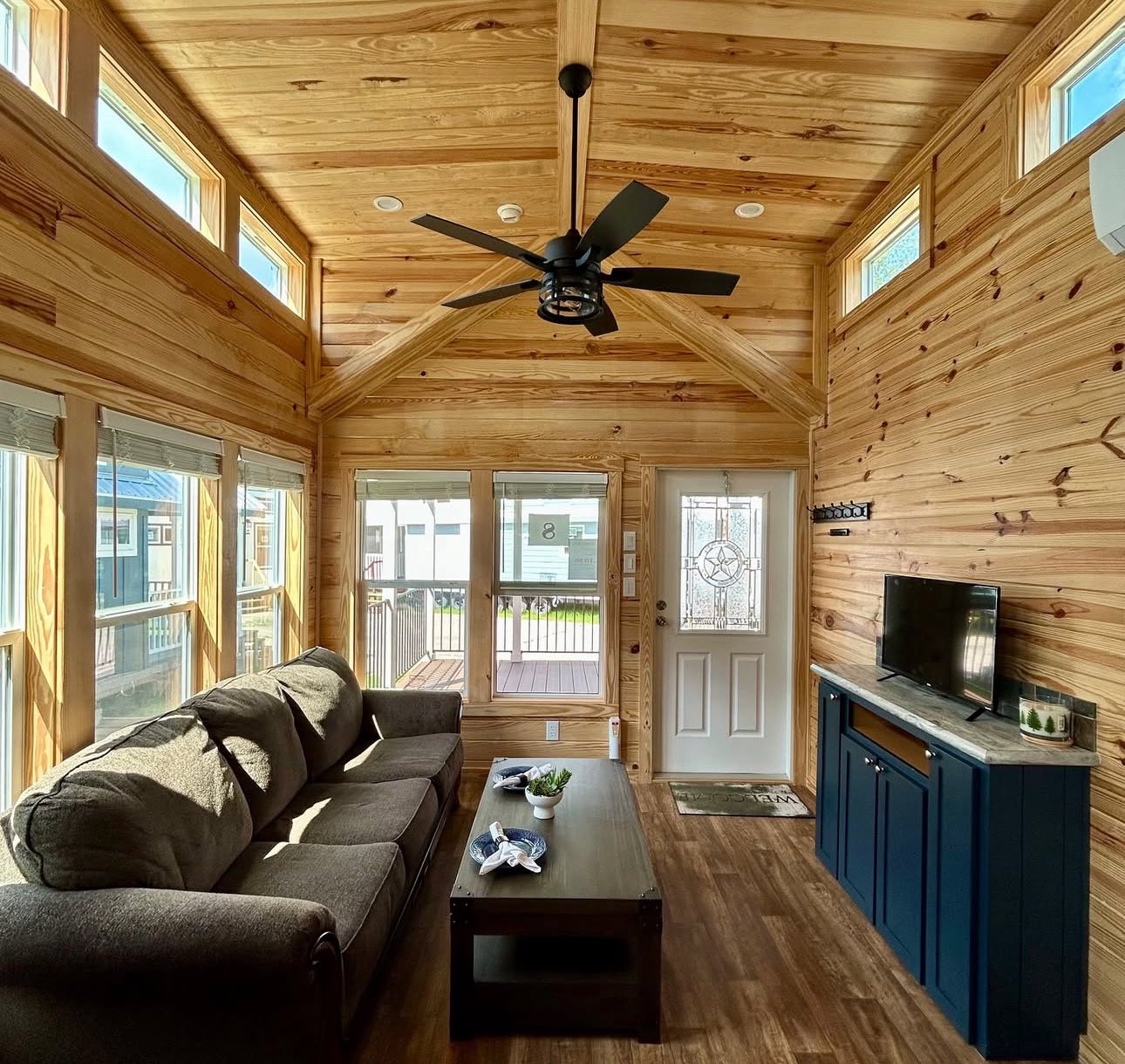 Cozy living room with wood-paneled walls and ceiling. Features large windows, a gray sofa, a wooden coffee table, TV, and ceiling fan. Bright, inviting space.