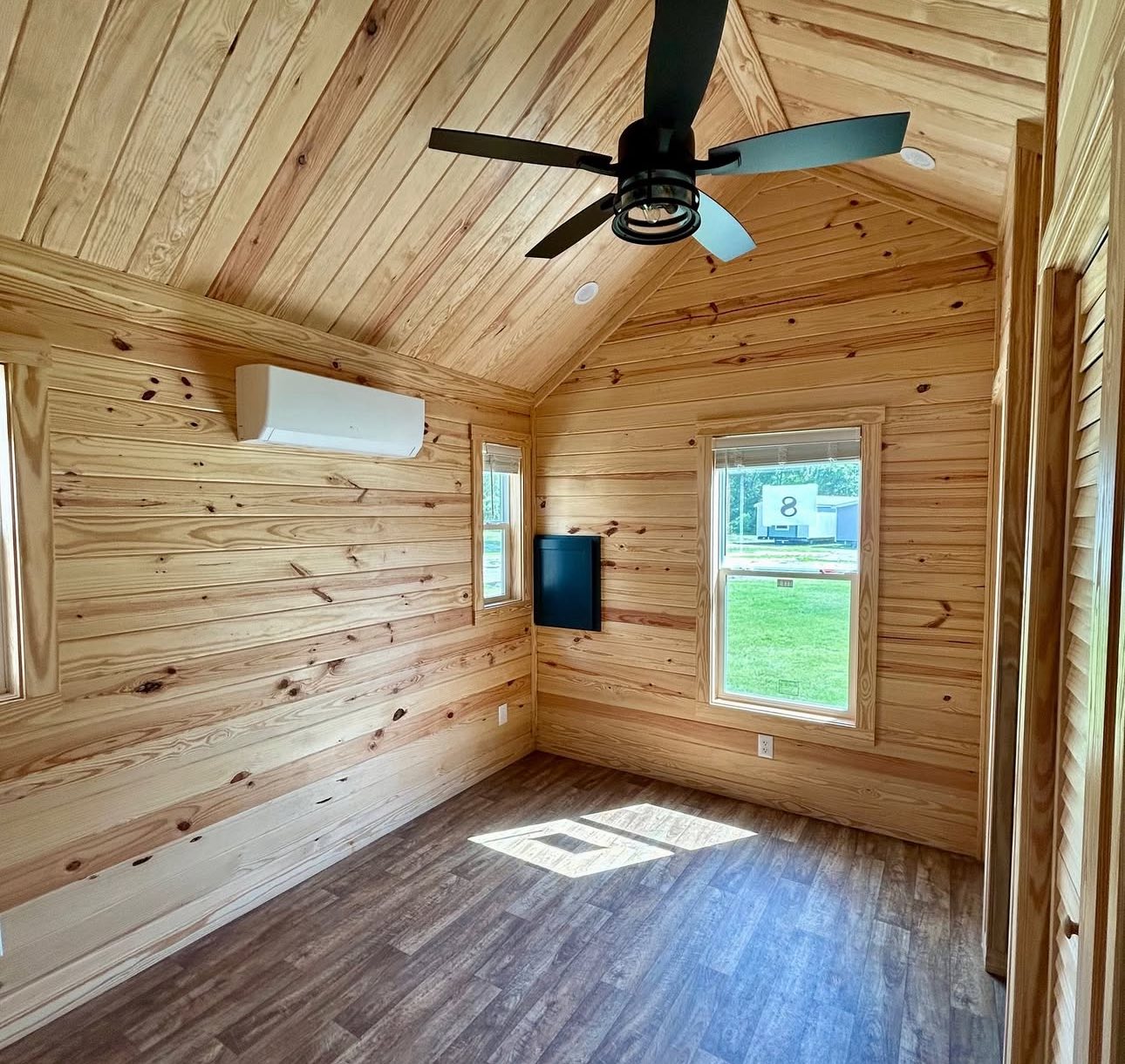 A cozy, sunlit room with wooden walls and ceiling. It features a black ceiling fan, air conditioner, mounted TV, and a window revealing a grassy lawn outside.