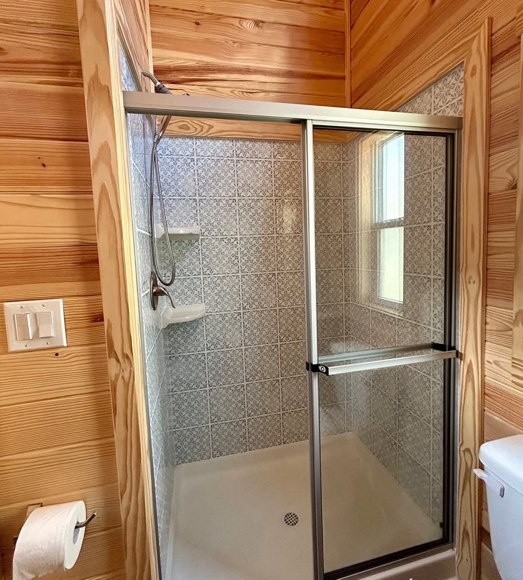 Wood-paneled bathroom with a glass-enclosed shower featuring patterned tiles and a window. Toilet paper holder and shower fixtures visible. Cozy and rustic.