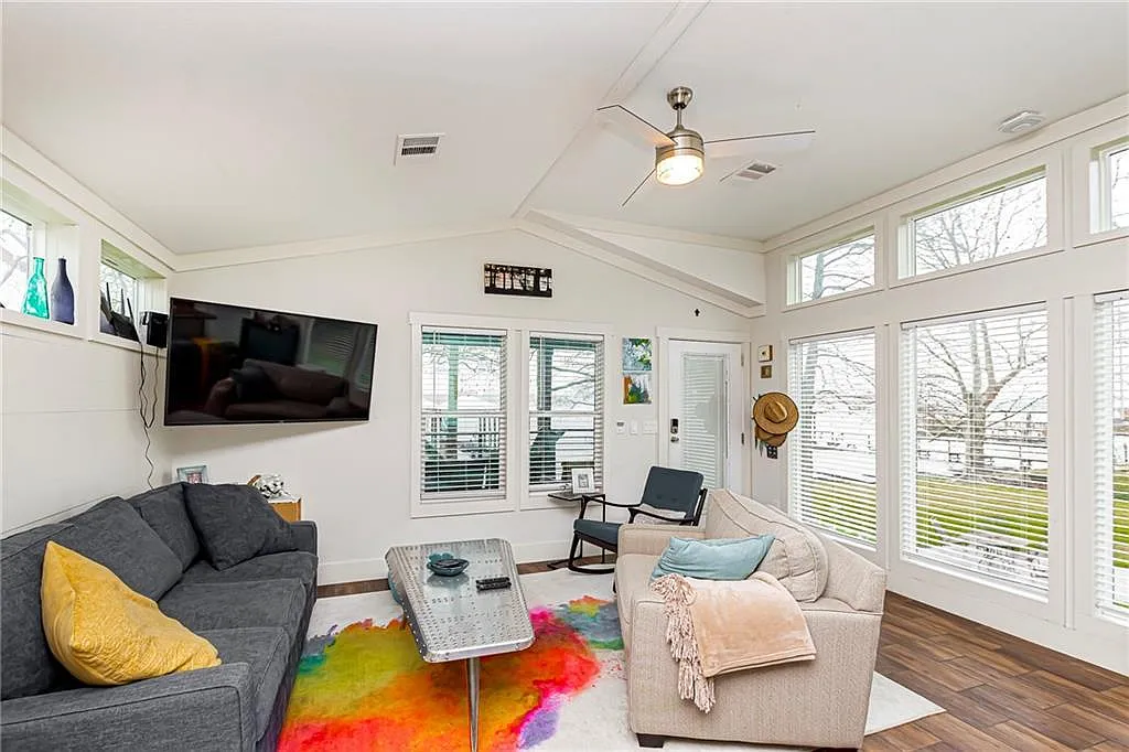 A bright living room with large windows and white walls. It features a gray sofa with a yellow pillow, an armchair, a colorful rug, and a wall-mounted TV. A ceiling fan hangs from the vaulted ceiling, creating a cozy, modern feel.