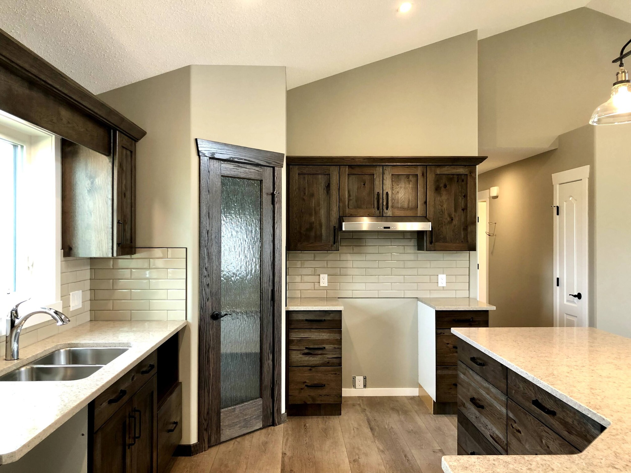 Modern kitchen with dark wood cabinets, light countertops, and stainless steel sink and range hood. Beige subway tiles and frosted pantry door. Bright, spacious tone.