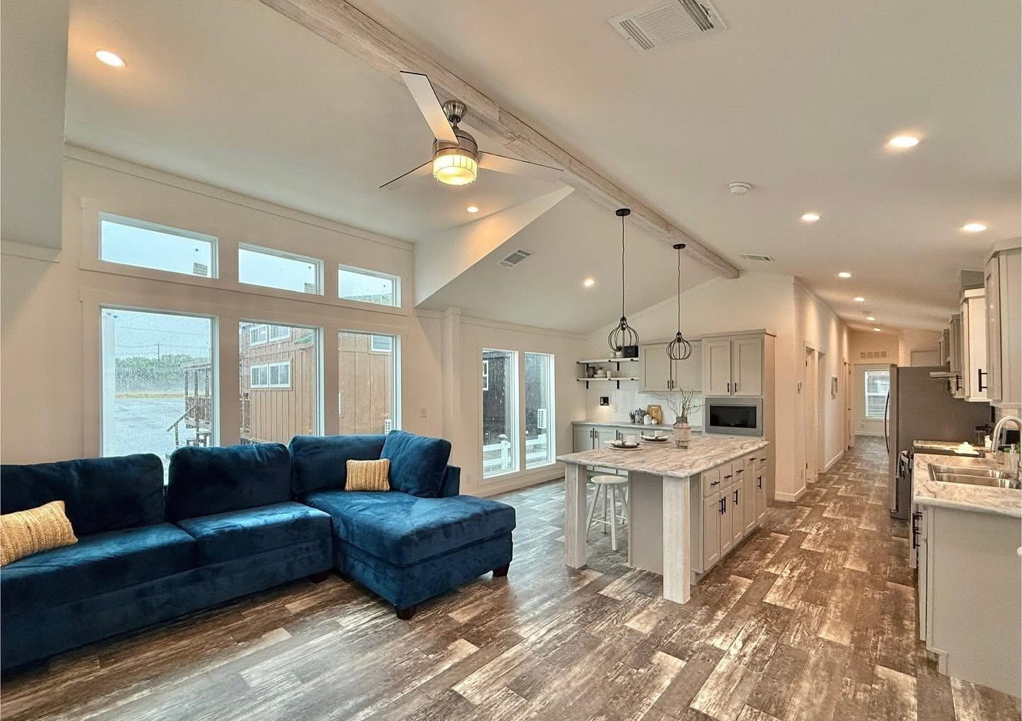 Spacious living area with blue sectional sofa, yellow pillows, and large windows. Modern kitchen features marble countertops and pendant lights. Cozy and inviting.