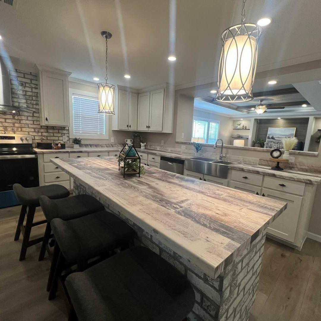 Modern kitchen with a rustic island, pendant lights, and black bar stools. Brick accents, white cabinets, and stainless steel appliances create a cozy, inviting vibe.