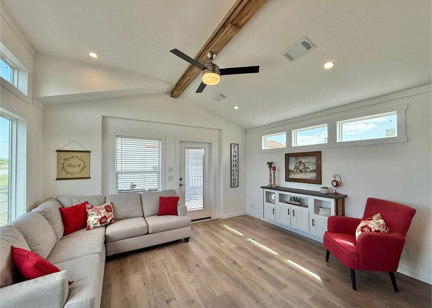 Bright, cozy living room with vaulted ceiling and wood beam. Features a beige sectional, red chair, pillows, farmhouse decor, and wood flooring.