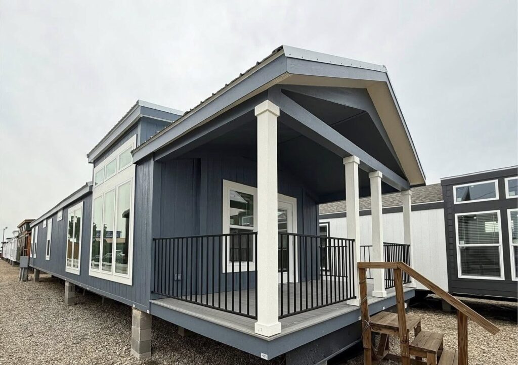 Modern blue manufactured home with a front porch, white trim, tall windows, and steps leading up. Set in a gravel lot, conveying a calm, neat appearance.