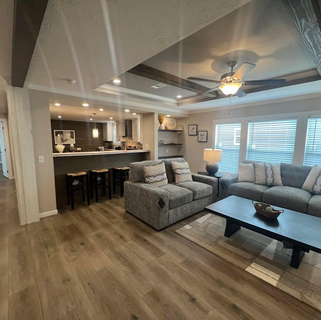 A cozy living room features a gray sectional sofa, cushioned armchairs, and a dark wooden coffee table. A ceiling fan spins above, soft lighting brightens the space from the kitchen bar area, which includes three bar stools. The laminate wood flooring complements the warm and inviting ambiance.