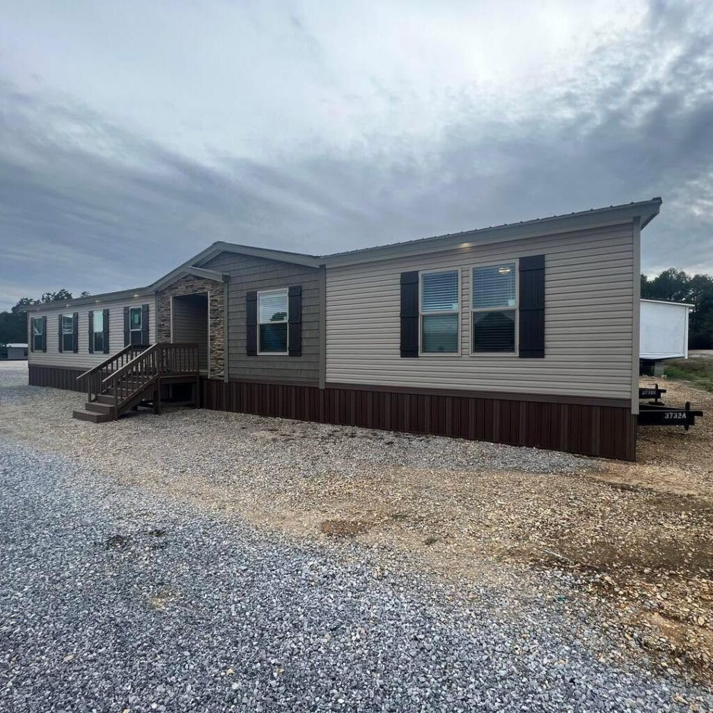 Single-story modular home with beige siding and dark shutters, set on a gravel lot under a cloudy sky; a small wooden porch is visible.