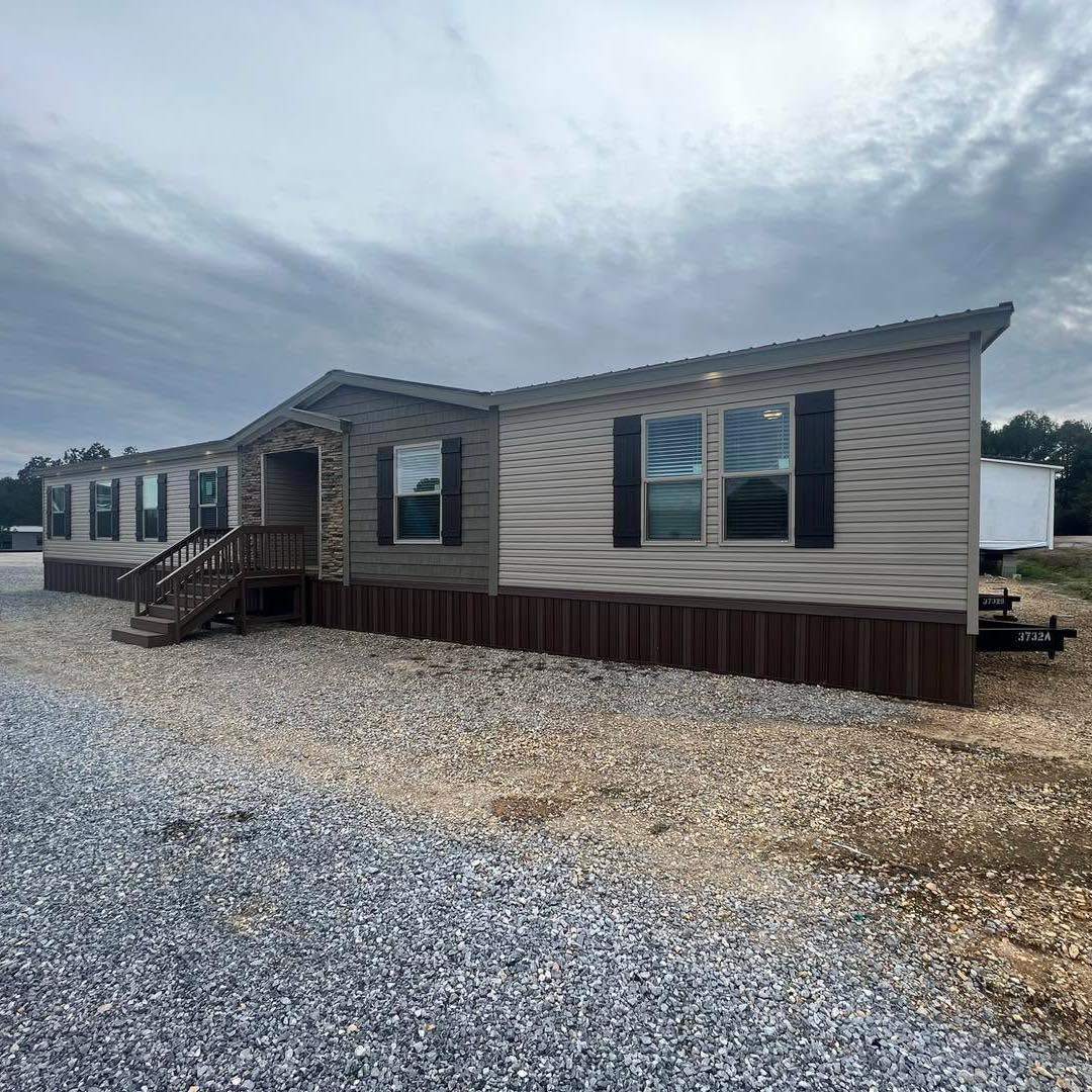 Single-story modular home with beige siding and dark shutters, set on a gravel lot under a cloudy sky; a small wooden porch is visible.