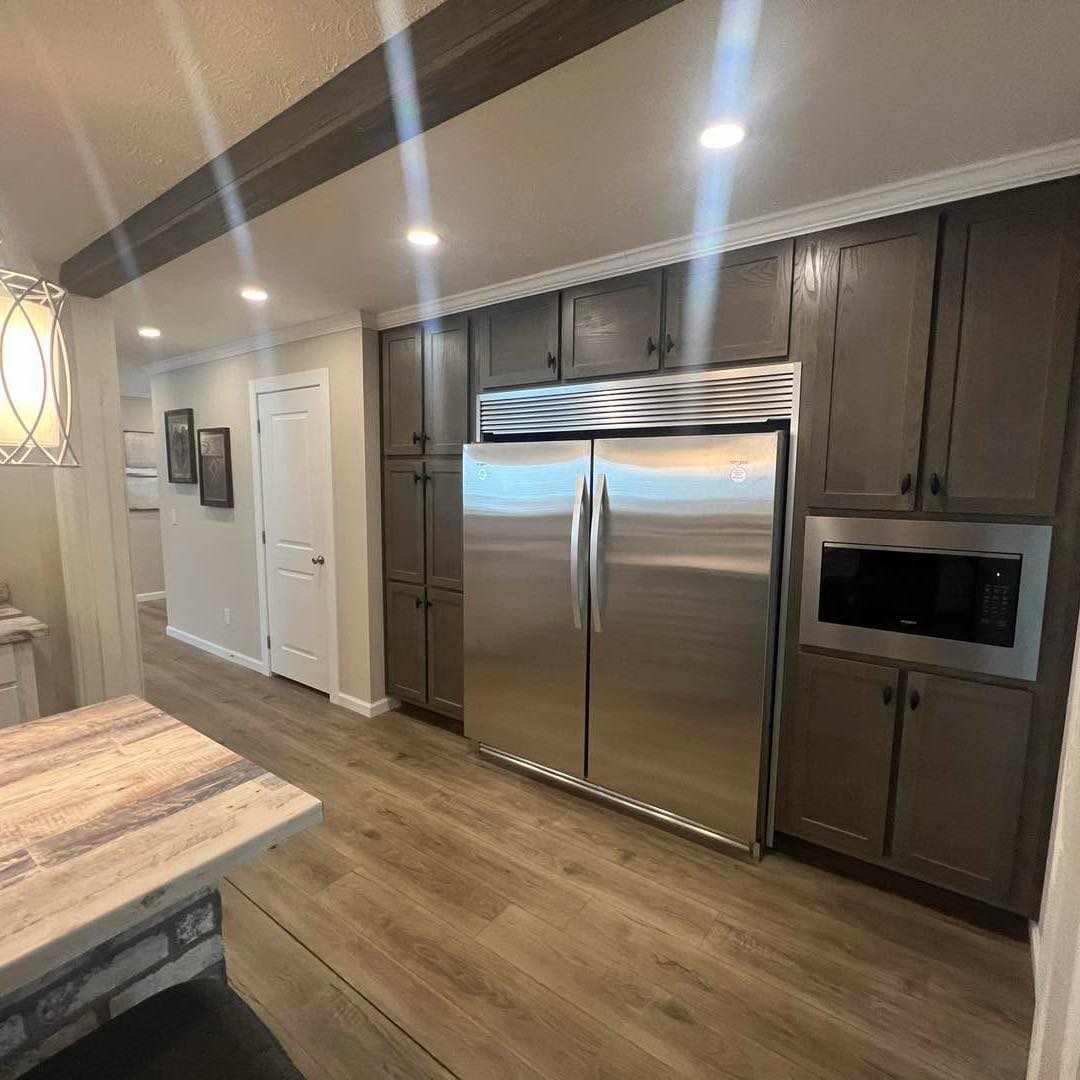 Modern kitchen with wood flooring and dark brown cabinets. A large stainless steel fridge is centered, flanked by cabinets and a microwave. Warm lighting creates a cozy ambiance.